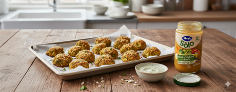 Bocaditos de brócoli dorados sobre papel vegetal en una bandeja, con un cuenco de salsa y un tarrito de verduras Hero Solo